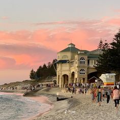 people walking on the beach at sunset near an old building with a pink sky in the background