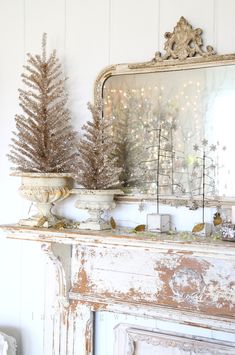 a white mantle with two potted trees sitting on it's side and a mirror behind it