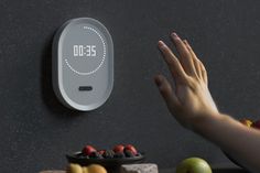 a person's hand reaching for an electronic clock on the wall next to fruit