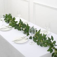 the table is set with white plates and silverware, greenery, and wine glasses