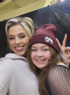 two women are posing for the camera in front of a black background and one is wearing a red beanie