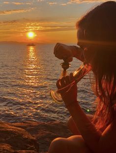 a woman drinking from a wine glass while sitting on the rocks by the ocean at sunset