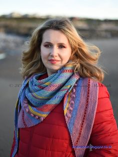 a woman standing on the beach wearing a red coat and blue scarf over her shoulders