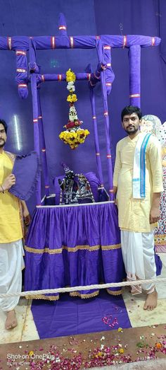 two men standing in front of a purple and yellow decorated stage with flowers on it