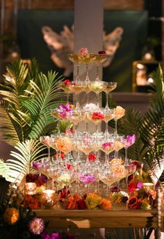 a tall tower filled with lots of wine glasses next to flowers and greenery in front of a statue