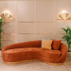 an orange curved couch sitting in front of two potted plants and gold fan lights