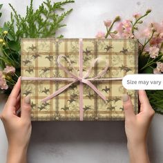 two hands holding a wrapped gift box with pink ribbon and bow on it, surrounded by flowers