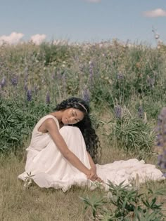 a woman in a white dress sitting on the ground next to purple flowers and grass