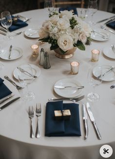 a white table with blue napkins, silverware and flowers in a gold vase