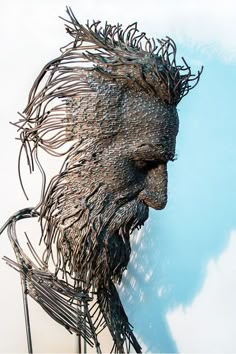 a metal sculpture of a man's face with hair blowing in the wind, against a blue sky