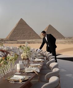 a man in a tuxedo standing next to a long table with plates and glasses on it