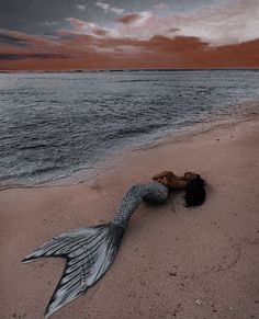 a mermaid tail laying on the beach next to the ocean at sunset with red clouds in the background