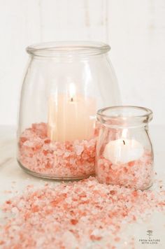 two glass jars filled with pink and white sand next to a lit candle on top of a table