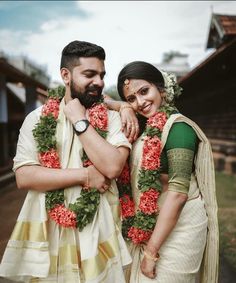 a man and woman standing next to each other in front of a building with flowers around their necks