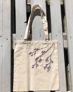 a tote bag with purple flowers on it hanging from a wooden fence, against a white background