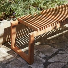 a wooden bench sitting on top of a stone floor next to bushes and flowers in the sun