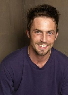 a close up of a person wearing a blue shirt and smiling at the camera with a green wall in the background