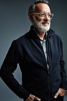 an older man with glasses and a beard standing in front of a gray background wearing a black jacket