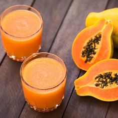 two glasses filled with orange juice next to some fruit on a wooden table and one is cut in half