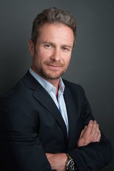 a man with his arms crossed wearing a suit and smiling at the camera while standing in front of a dark background