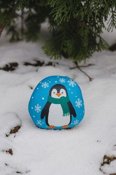 a rock with a penguin painted on it sitting in the snow next to a tree