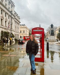 a woman standing in front of a red phone booth on a wet street with buildings behind her