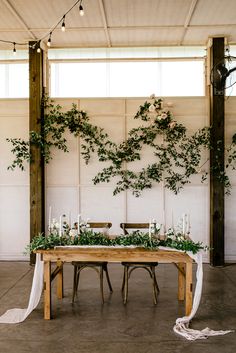 a wooden table topped with greenery next to a wall covered in white cloths