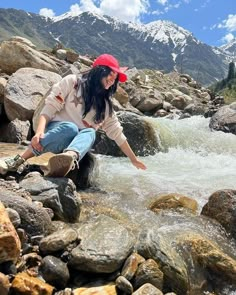 a woman sitting on rocks in the middle of a river with mountains in the background