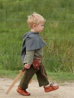 a little boy walking across a dirt road holding a wooden stick and wearing a cape