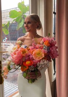 a woman in a wedding dress holding a bouquet of pink and orange flowers by a window