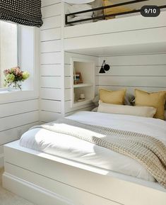 a white bed sitting under a window next to a wooden shelf filled with flowers and books