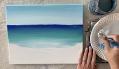 a person is painting an ocean scene with blue and white colors on paper plates, while another hand holds a paintbrush