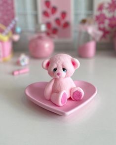 a small pink teddy bear sitting on top of a heart shaped plate with other toys in the background