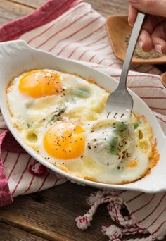 two fried eggs in a white dish on a wooden table with a fork and spoon