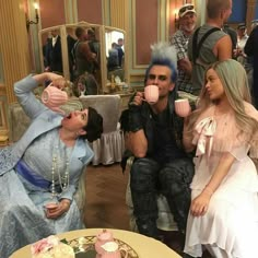 three people sitting at a table with cupcakes in front of them and one woman drinking from a mug