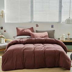 a bed with red sheets and pillows in a room next to a white window,