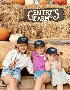 three young children sitting on a wooden bench in front of hay bales and pumpkins