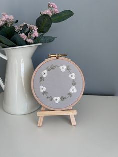 a white vase with pink flowers next to a gray and white cross stitched flower arrangement