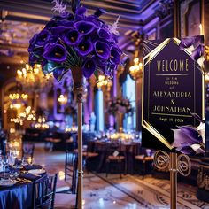 purple flowers are in a tall vase next to a welcome sign at a wedding reception