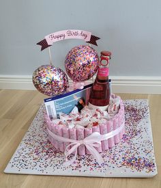 a pink birthday cake with confetti, balloons and wine bottle on the table