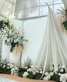 an arrangement of flowers and greenery is displayed in front of a large white wall