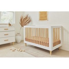 a baby crib in a white room next to a dresser and window with palm leaves on it