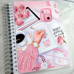 a spiral notebook with pink flowers and writing on it, next to a pen and camera