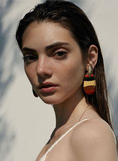 a woman with long hair wearing earrings and looking at the camera while standing in front of a white wall