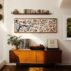 a wooden cabinet with books and plants on it in front of a wall mounted art piece