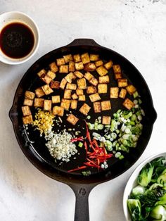 an iron skillet filled with tofu and vegetables next to two bowls of sauce