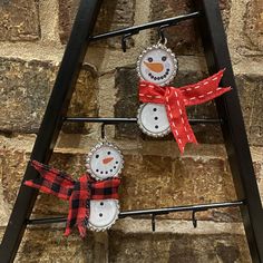 three snowman bottle cap ornaments hanging from a ladder on a brick wall with a red bow