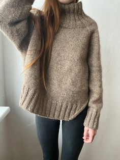 a young woman wearing a brown sweater and black leggings stands in front of a white wall