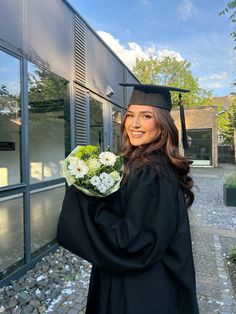 a woman wearing a graduation gown and holding a bouquet of flowers in front of a building