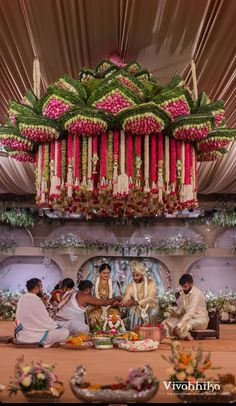 a group of people sitting around a cake on top of a table with flowers hanging from the ceiling